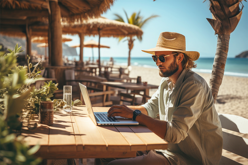 Man wearing hat & sunglasses working with laptop on table at beach with palmtrees and bamboo shelters.