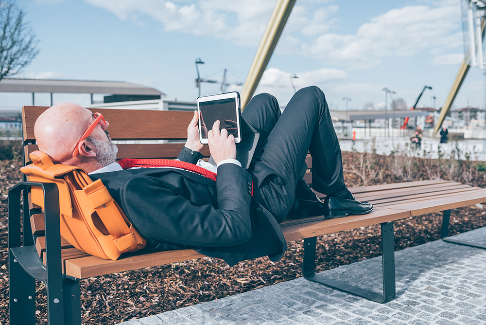 Businessman in suit at work site laying on park bench with tablet working.