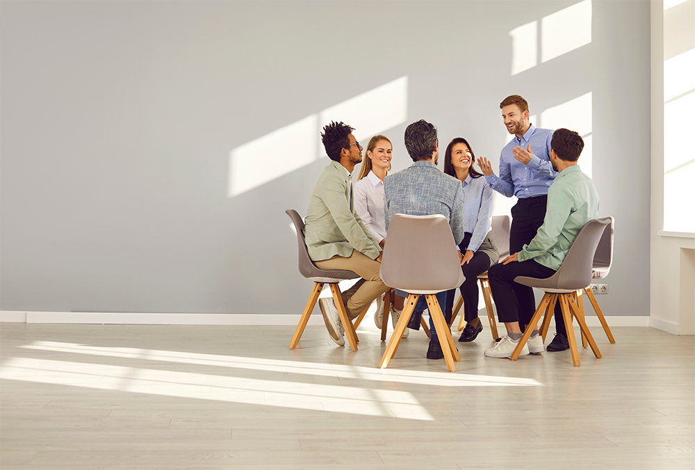 Group of employees huddled in circle with chairs at clean office with motivating person standing.