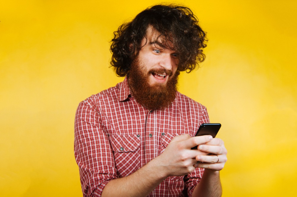 Surprised young bearded man using smart phone. Surprised man with beard reading or writing a message. Handsome man with curly hair and beard wearing checkered shirt..