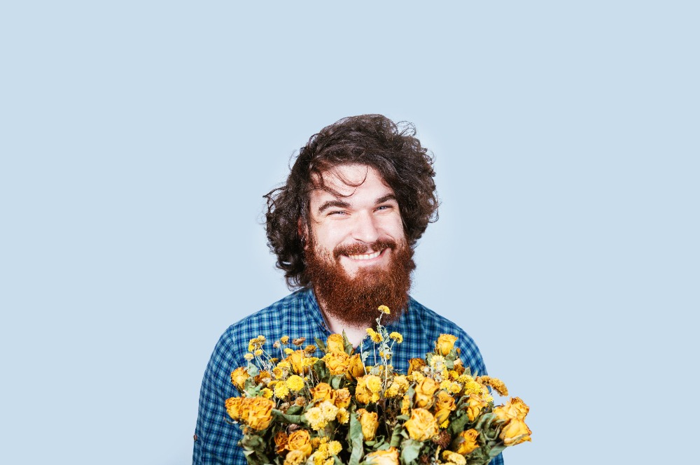 Smiling happy bearded man with bunch of dry yellow roses on white background. Curly hair. Love peace and flowers. Man with flowers looking at camera.