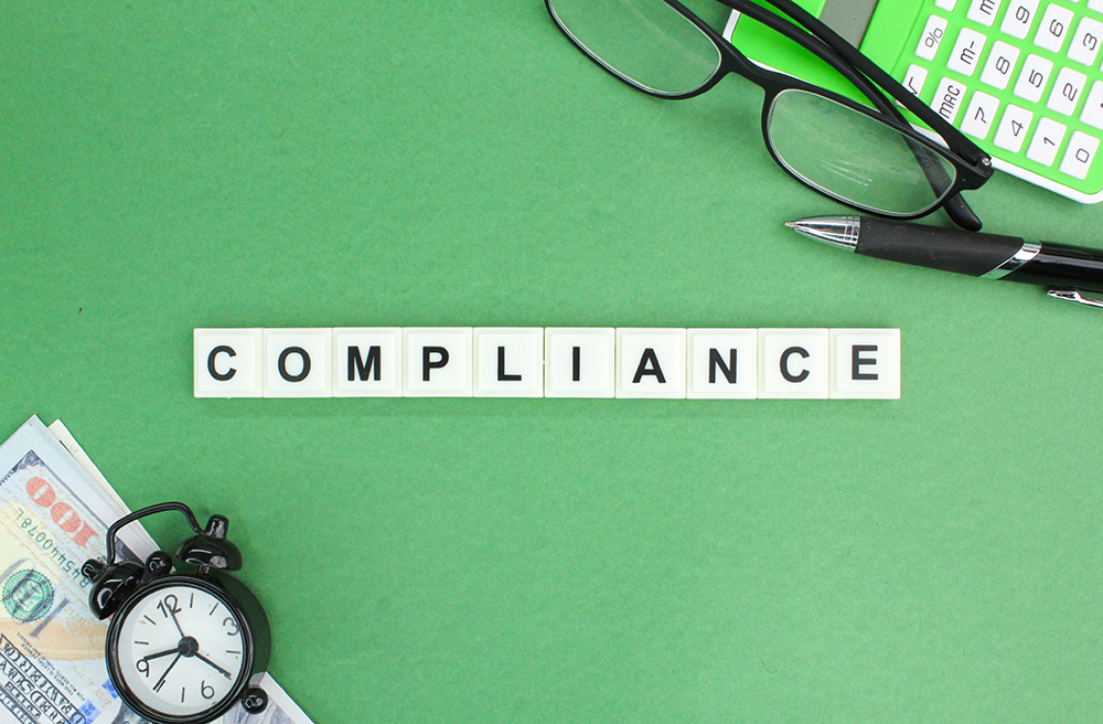 Scrabble pieces spelling the word compliance.  Other items like glasses, alarm clock and calculator on desk.