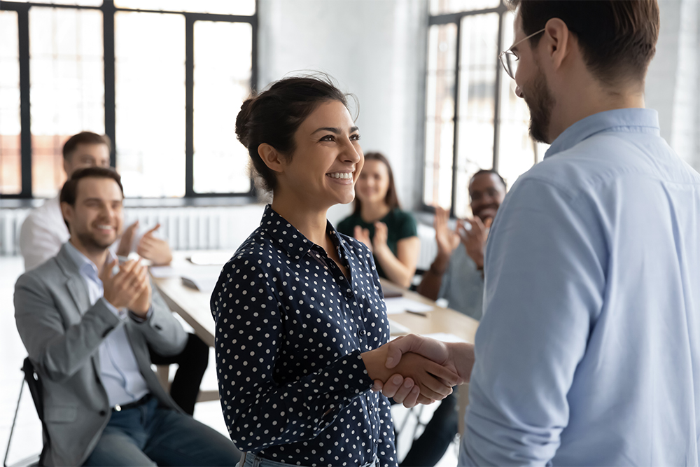 Happy proud excited Indian ethnicity employee get promotion receive praises from boss and cheering and congratulations from diverse staff members shake hands with chief. Recognition of success