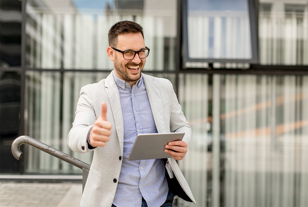 Happy businessman with tablet outside office giving thumbs up.