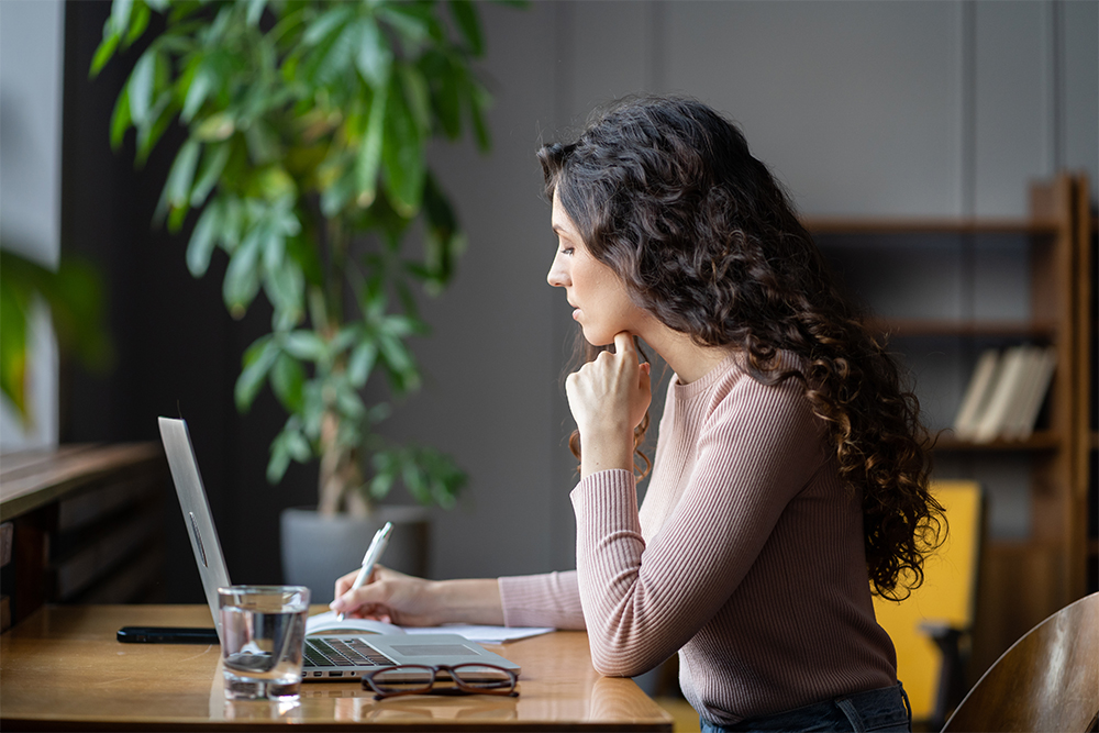 woman writing notes in front of laptop at desk in modern office with plants