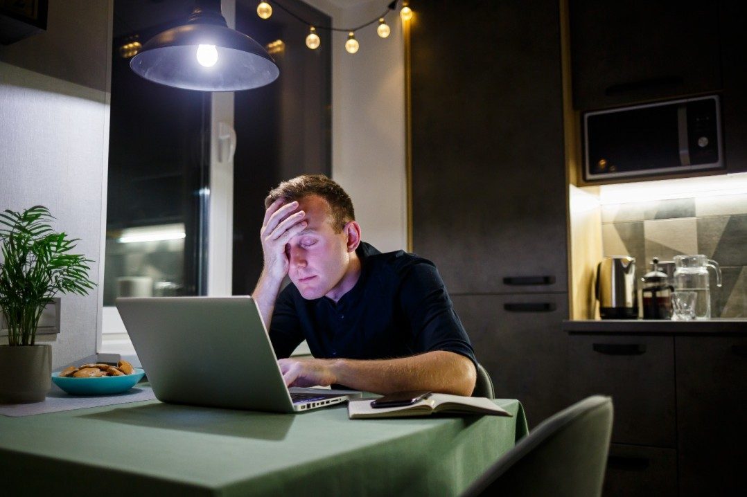 Tired exhausted overworked man siting at office desk with laptop lost data and has head in his hands