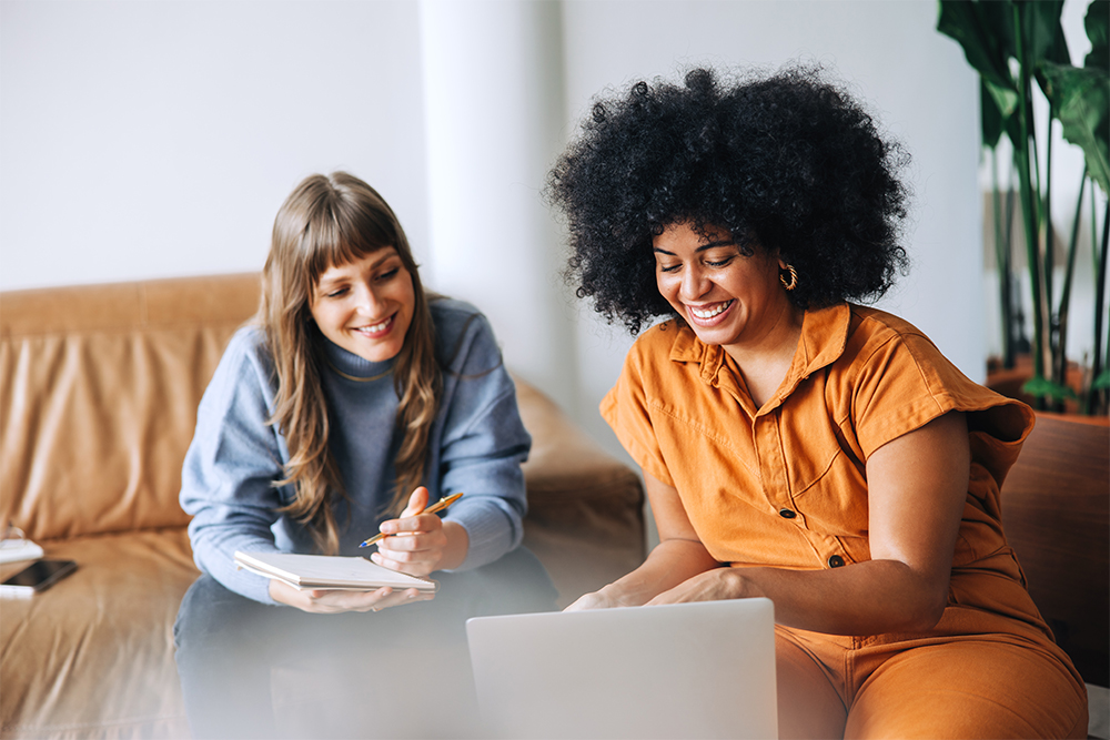 Happy young business women learning and sharing a laptop together in a light filled office on a lounge.