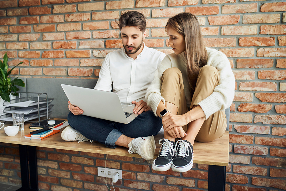 Two young people sitting on office desk using laptop and talking.