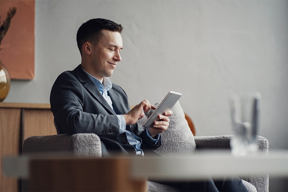 Man in a business suit sitting in a modern office, using a tablet, smiling.